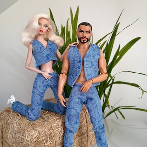 Puede incluir: Dos muñecos de moda, un hombre y una mujer, vestidos con conjuntos de mezclilla azul a juego con un diseño de cachemira. La muñeca femenina está arrodillada sobre una paca de heno, mientras que el muñeco masculino está a su lado. El fondo incluye plantas verdes y una pared blanca.