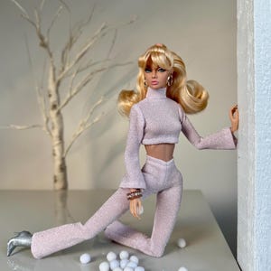 Pu&ograve; includere: Una bambola alla moda indossa un completo a due pezzi rosa chiaro e strutturato, con un top corto a collo alto e pantaloni svasati. La bambola ha i capelli biondi, orecchini a cerchio dorati e scarpe argentate. Piccole palline bianche sono sparse.