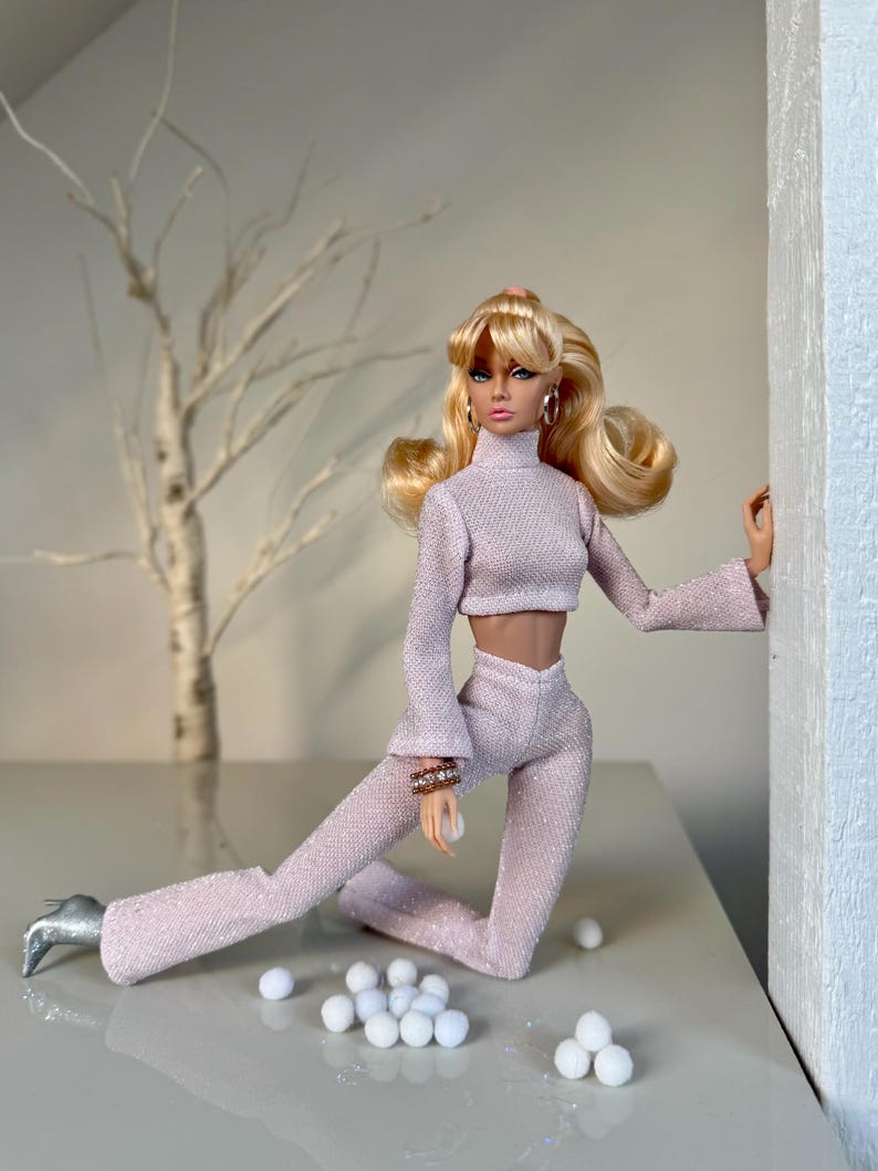 Pu&ograve; includere: Una bambola alla moda indossa un completo a due pezzi rosa chiaro e strutturato, con un top corto a collo alto e pantaloni svasati. La bambola ha i capelli biondi, orecchini a cerchio dorati e scarpe argentate. Piccole palline bianche sono sparse.