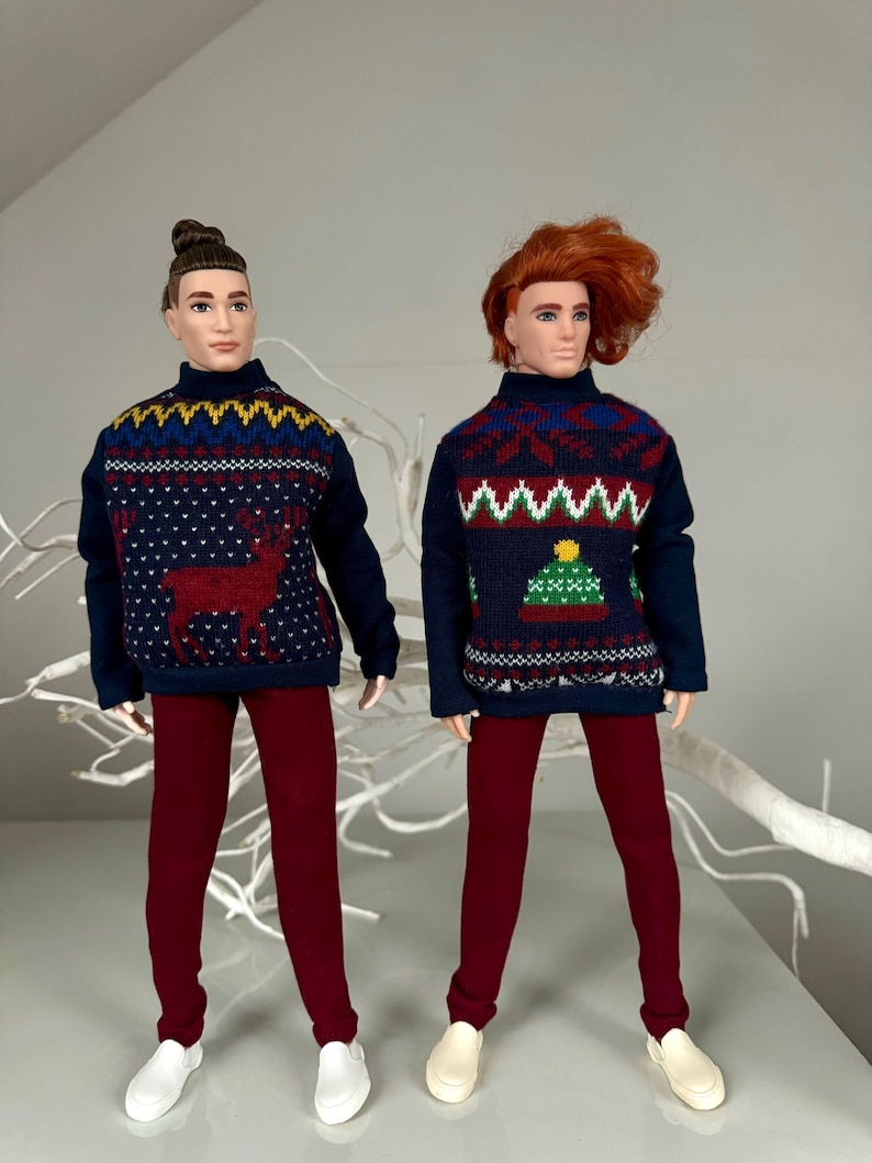 May include: Two dolls wearing festive holiday sweaters with different patterns. One sweater features a reindeer, while the other has a Christmas tree design. Both dolls are wearing maroon pants and white shoes.