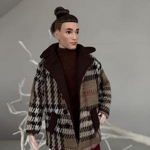 Könnte beinhalten: Eine Puppe trägt einen braunen und weißen karierten Mantel mit einem braunen Rollkragenpullover und einer maroonfarbenen Hose. Die Puppe hat braunes Haar, das zu einem Knoten gebunden ist.