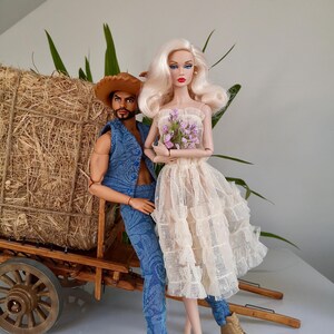 May include: Two fashion dolls, a man and a woman, are posed together. The man doll wears a denim vest, jeans, and a cowboy hat. The woman doll wears a cream-colored dress and holds a bouquet of purple flowers. They stand near a wooden cart.