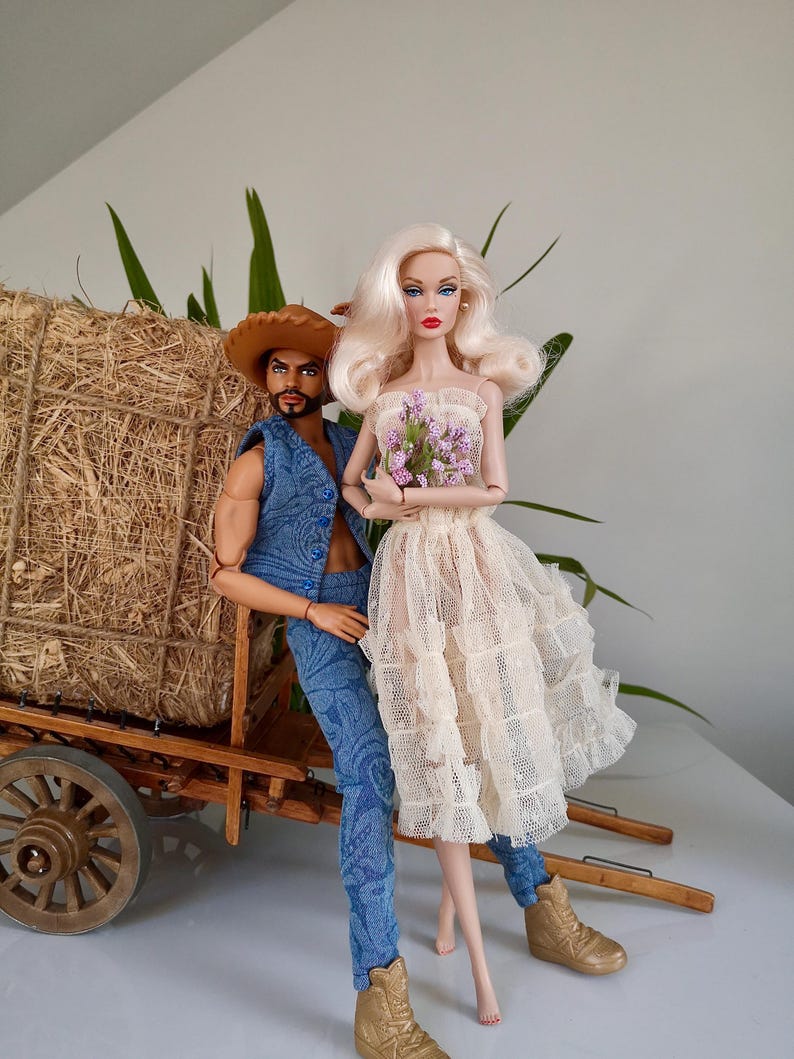 May include: Two fashion dolls, a man and a woman, are posed together. The man doll wears a denim vest, jeans, and a cowboy hat. The woman doll wears a cream-colored dress and holds a bouquet of purple flowers. They stand near a wooden cart.