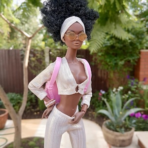 May include: A fashion doll with dark brown skin, wearing a white and gold outfit, a pink headband, and round sunglasses. The doll is carrying a pink backpack.