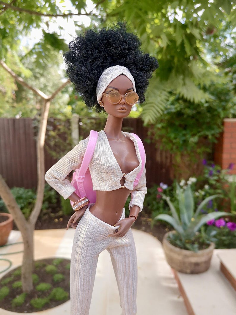 May include: A fashion doll with dark brown skin, wearing a white and gold outfit, a pink headband, and round sunglasses. The doll is carrying a pink backpack.