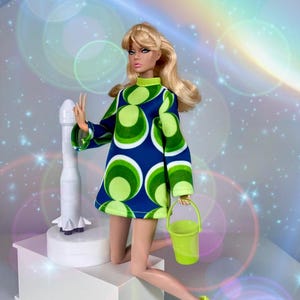 May include: A blonde fashion doll wearing a blue and green circular patterned shift dress with long sleeves and a high neckline. The doll holds a lime green bucket in one hand and wears matching lime green ankle boots, positioned next to a white rocket.