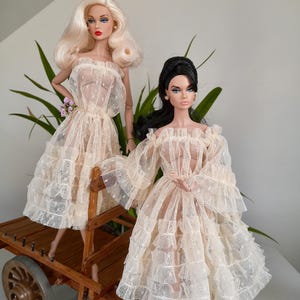 May include: Two fashion dolls in cream-colored lace dresses. One doll has blonde hair, the other has black hair. They are posed on a wooden cart with a single wheel. The dolls are wearing off-the-shoulder dresses with ruffled sleeves and skirts.