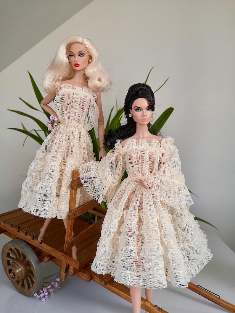 May include: Two fashion dolls in cream-colored lace dresses. One doll has blonde hair, the other has black hair. They are posed on a wooden cart with a single wheel. The dolls are wearing off-the-shoulder dresses with ruffled sleeves and skirts.