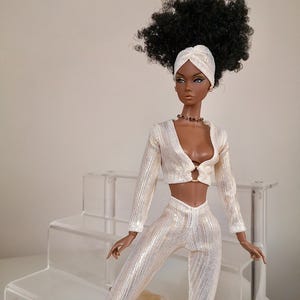May include: A fashion doll with dark brown skin, black curly hair, and a white turban. She is wearing a white, sparkly, two-piece outfit with a long-sleeved top and trousers. The doll is sitting on a white, tiered display stand.