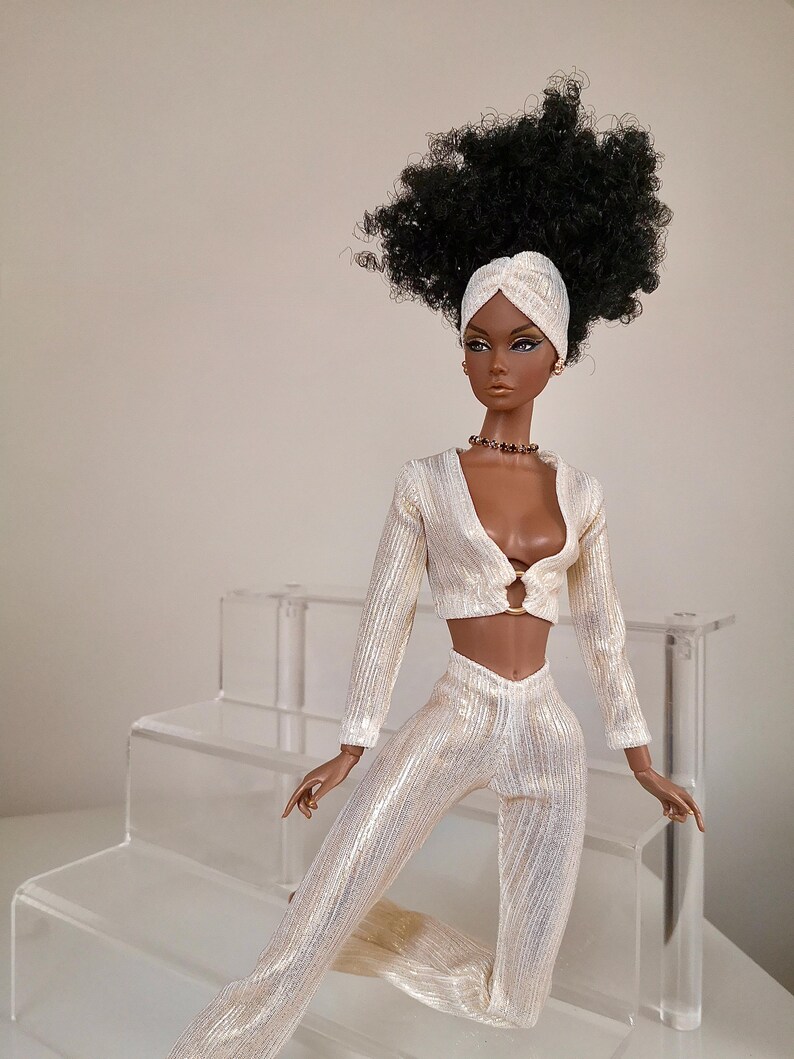 May include: A fashion doll with dark brown skin, black curly hair, and a white turban. She is wearing a white, sparkly, two-piece outfit with a long-sleeved top and trousers. The doll is sitting on a white, tiered display stand.
