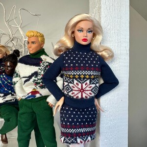 May include: Three dolls wearing festive holiday sweaters. The doll in the center is a blonde doll wearing a blue and red sweater with a snowflake design. The doll on the left is wearing a white and green sweater with a Christmas tree design. The doll on the right is wearing a green sweater with a Christmas tree design.