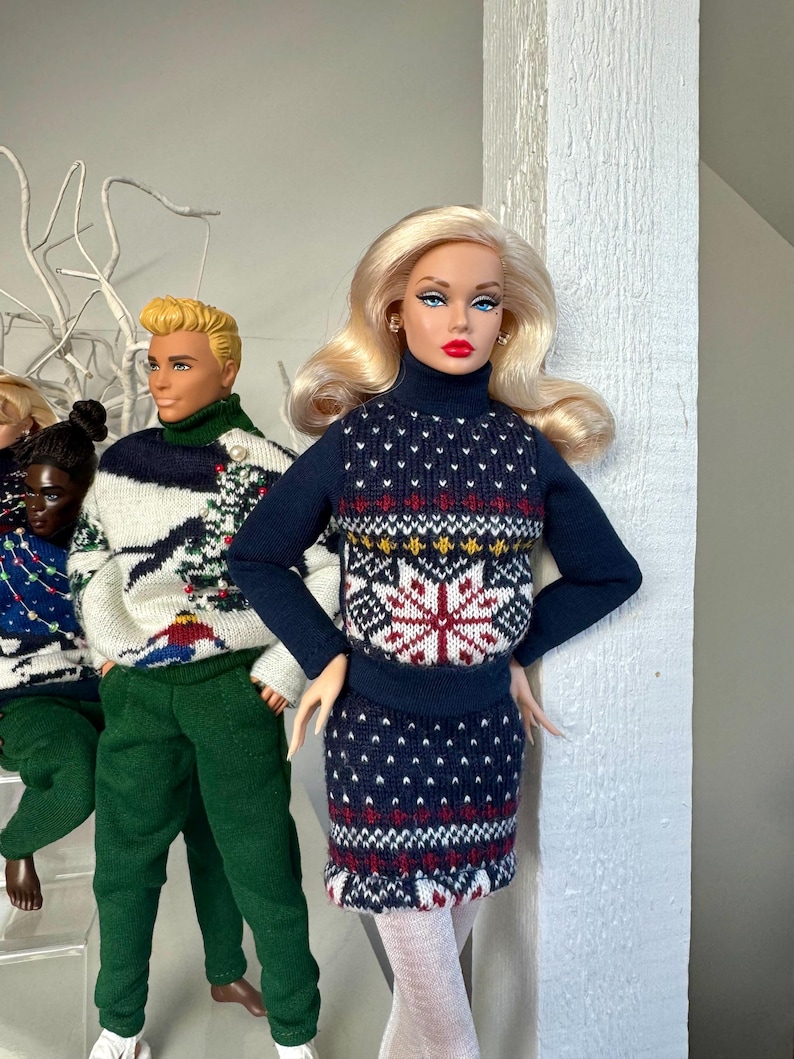May include: Three dolls wearing festive holiday sweaters. The doll in the center is a blonde doll wearing a blue and red sweater with a snowflake design. The doll on the left is wearing a white and green sweater with a Christmas tree design. The doll on the right is wearing a green sweater with a Christmas tree design.