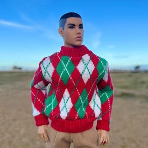 May include: A doll wearing a red, green, and white argyle sweater with a red turtleneck and cuffs. The doll is also wearing tan trousers. The background is a blue sky.