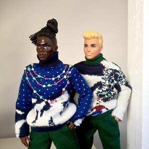 May include: Two dolls wearing festive holiday sweaters. One doll has dark skin and a braided bun, wearing a blue and white sweater with a string of colorful lights. The other doll has blonde hair and is wearing a green and white sweater with a Christmas tree design.