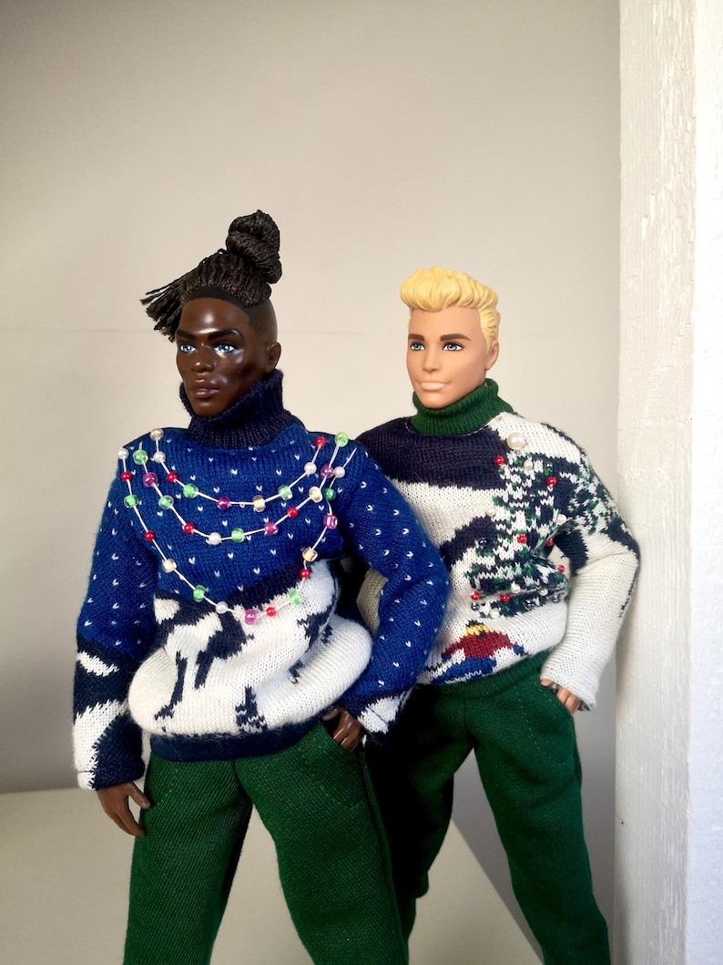 May include: Two dolls wearing festive holiday sweaters. One doll has dark skin and a braided bun, wearing a blue and white sweater with a string of colorful lights. The other doll has blonde hair and is wearing a green and white sweater with a Christmas tree design.