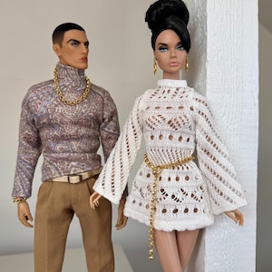 Op de afbeelding: Twee poppen, een met een gepatroneerde trui en een bruine broek, de andere draagt een witte gehaakte jurk met een gouden kettingriem.