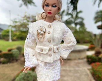May include: A fashion doll wearing a white tweed skirt suit with gold buttons and a flower brooch. The doll has blonde hair, blue eyes, and red lipstick. Gold hoop earrings complete the ensemble. The doll is standing on a paved walkway.