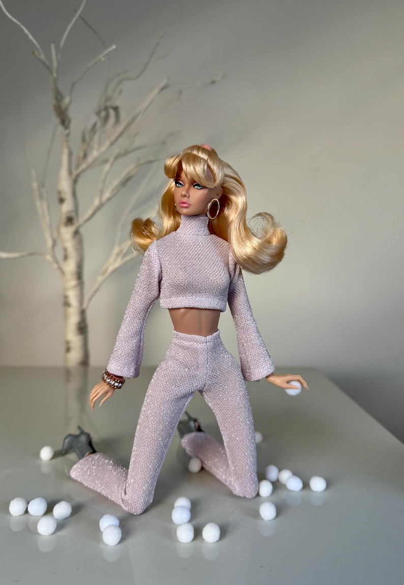 Pu&ograve; includere: Bambola alla moda bionda con orecchini a cerchio, indossa un top corto rosa chiaro strutturato e pantaloni abbinati. La bambola &egrave; in posa con piccole palline bianche sparse. Un albero bianco decorativo &egrave; sullo sfondo.