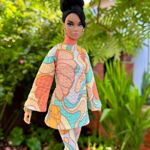 May include: A fashion doll wearing a colorful, patterned outfit. The ensemble includes a long-sleeved top and matching pants in shades of orange, yellow, and blue. The doll has black hair styled in a high bun and is wearing gold hoop earrings.