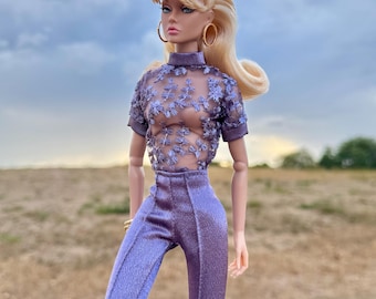 Può includere: Una bambola alla moda con capelli biondi raccolti in una coda di cavallo alta, indossa orecchini a cerchio dorati, un top trasparente viola a maniche corte con intricati ricami floreali e pantaloni viola abbinati con finitura satinata.