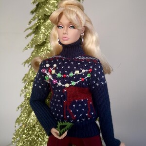 May include: A doll wearing a blue knitted sweater with a reindeer and Christmas lights design. The sweater has white and red accents. The doll has blonde hair and is wearing red pants.