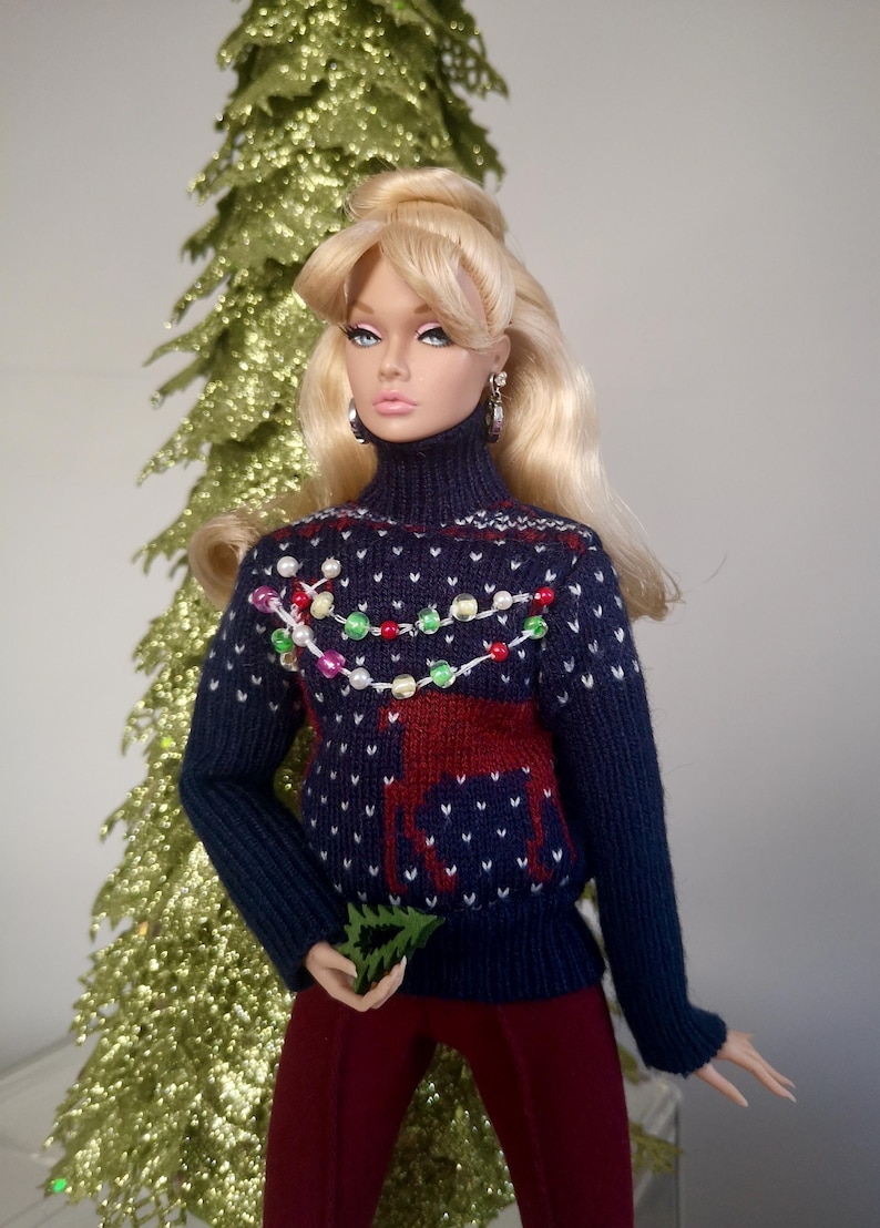 May include: A doll wearing a blue knitted sweater with a reindeer and Christmas lights design. The sweater has white and red accents. The doll has blonde hair and is wearing red pants.