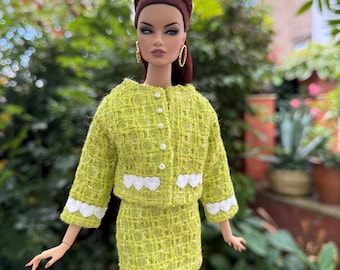 May include: A fashion doll wearing a lime green tweed skirt suit. The jacket has white heart-shaped trim and pearl buttons. The doll has brown hair and gold hoop earrings. The background is a blurred outdoor scene with greenery.