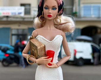 May include: A doll wearing a white dress, a black headscarf with floral print, and gold hoop earrings. The doll is holding a red cup and a small, gold-coloured book.
