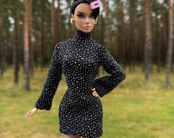 Può includere: Un vestito con motivo bianco e nero con collo alto e maniche lunghe. Il vestito è indossato da una bambola con i capelli neri raccolti in un alto chignon e una forcina rosa. La bambola è in piedi in un ambiente boschivo.