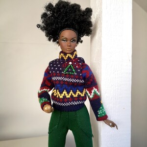 May include: A fashion doll with dark brown skin, wearing a dark blue, red, green, and yellow patterned sweater with a green and white triangle pin. The doll has dark brown eyes, black hair, and is wearing green pants.