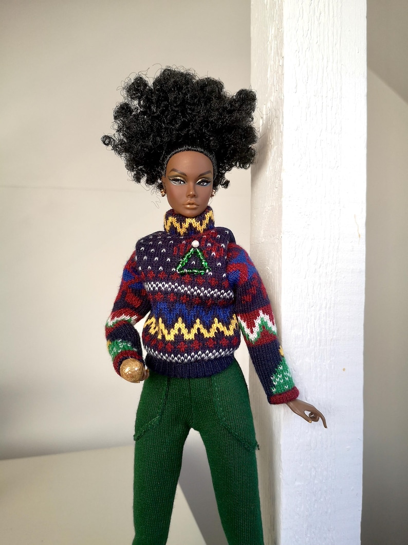 May include: A fashion doll with dark brown skin, wearing a dark blue, red, green, and yellow patterned sweater with a green and white triangle pin. The doll has dark brown eyes, black hair, and is wearing green pants.
