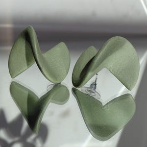 Può includere: Un paio di orecchini a lobo color verde salvia caratterizzati da una forma scultorea ritorta e ondulata. Gli orecchini hanno una finitura opaca e includono farfalline in plastica trasparente fissate ai perni metallici.