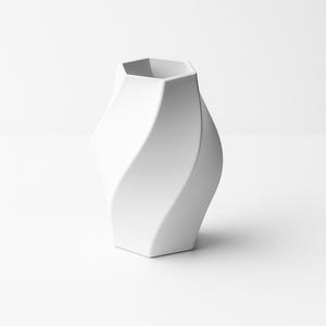 May include: A white ceramic vase with a unique, twisted design. The vase has a hexagonal opening and a smooth, matte finish. The overall shape is a modern, sculptural form, suitable for home decor.