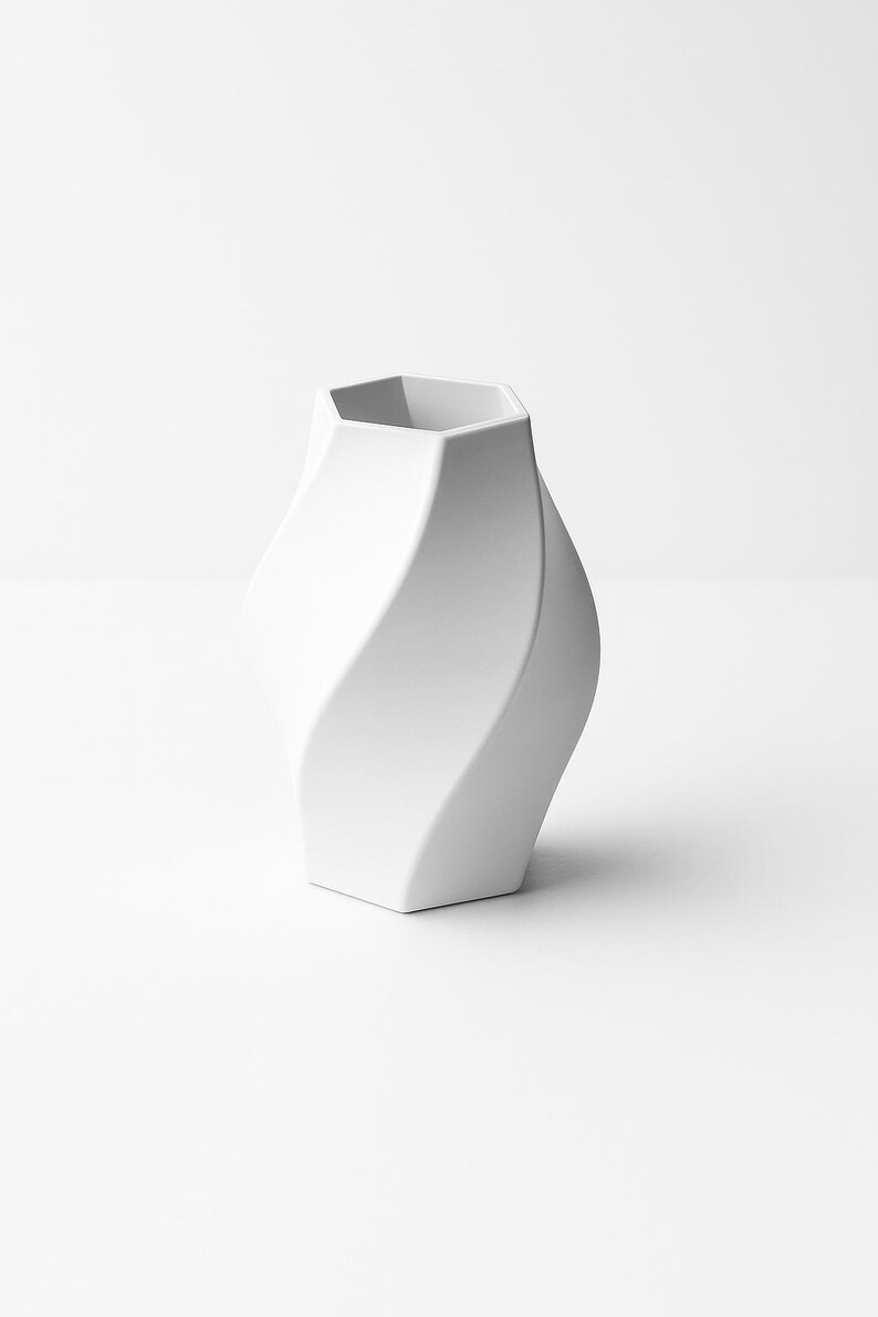 May include: A white ceramic vase with a unique, twisted design. The vase has a hexagonal opening and a smooth, matte finish. The overall shape is a modern, sculptural form, suitable for home decor.