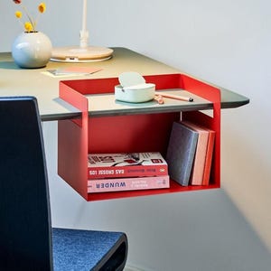 May include: A red metal desk organizer with two shelves, holding books and stationery. The top shelf has a small container and a pencil. The organizer is attached to a desk with a green surface.