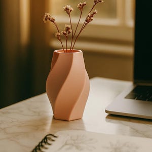 May include: A peach-colored, twisted design vase holding dried flowers. The vase sits on a marble surface, with a notebook and laptop in the background. The vase has a unique, modern shape.