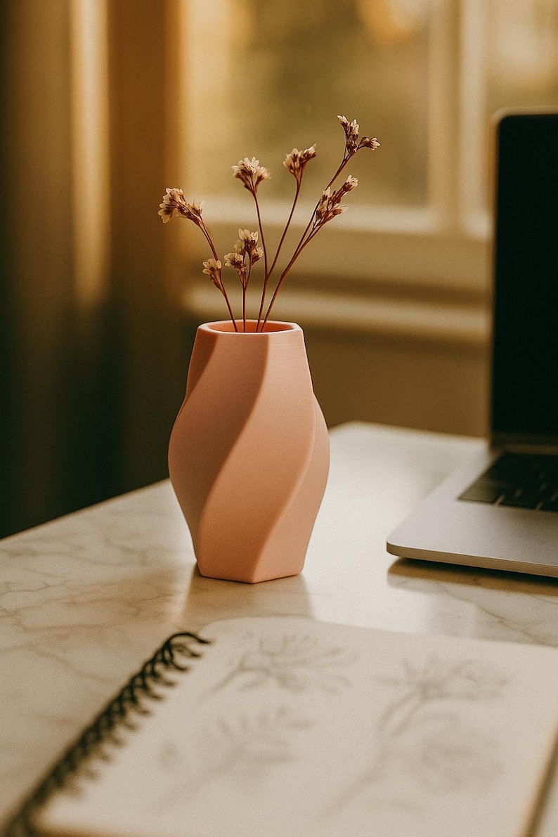 May include: A peach-colored, twisted design vase holding dried flowers. The vase sits on a marble surface, with a notebook and laptop in the background. The vase has a unique, modern shape.