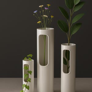 Puede incluir: Tres jarrones cilíndricos blancos de diferentes alturas, cada uno con un recorte rectangular. Los jarrones contienen vegetación y flores. El fondo es gris oscuro y la superficie es gris claro.