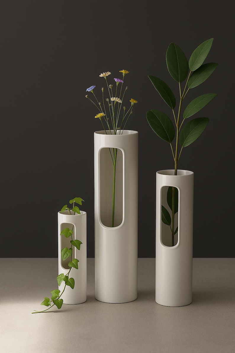 Op de afbeelding: Drie witte cilindervazen van verschillende hoogtes, elk met een rechthoekige uitsparing. De vazen bevatten groen en bloemen. De achtergrond is donkergrijs en het oppervlak is lichtgrijs.
