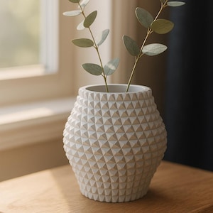 Pode incluir: Um vaso branco texturizado com um padrão geométrico, segurando dois ramos de folhas verdes de eucalipto. O vaso está sobre uma mesa de madeira perto de uma janela, criando uma estética natural e minimalista.