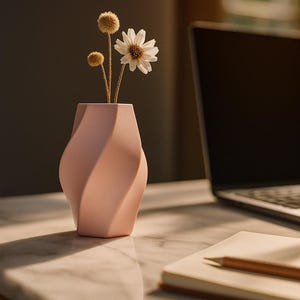 May include: A pale pink, contemporary vase with a twisted design holds dried flowers and a single white daisy. The vase sits on a marble surface next to a laptop and a notebook with a pencil.