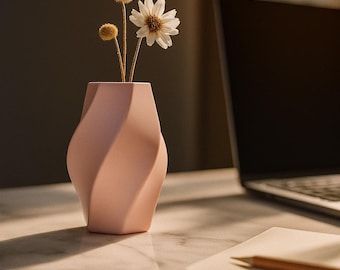 Könnte beinhalten: Eine hellrosa, moderne Vase mit gedrehtem Design hält getrocknete Blumen und eine einzelne weiße Gänseblümchen. Die Vase steht auf einer Marmoroberfläche neben einem Laptop und einem Notizbuch mit einem Bleistift.