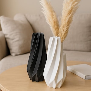 Puede incluir: Dos jarrones decorativos modernos, uno negro y otro blanco, con un diseño geométrico retorcido. El jarrón blanco contiene hierba de la pampa seca. Los jarrones están sobre una mesa de madera clara, con un sofá de tonos neutros al fondo.