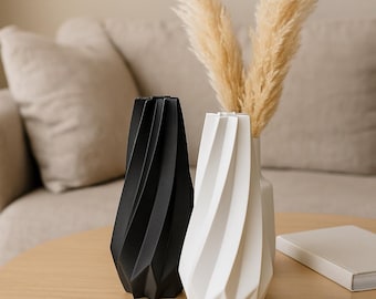 Könnte beinhalten: Zwei moderne Dekovasen, einer schwarz und einer weiß, mit einem geometrischen, gedrehten Design. Die weiße Vase enthält getrocknetes Pampasgras. Die Vasen stehen auf einem hellen Holztisch, im Hintergrund ein Sofa in neutralen Tönen.