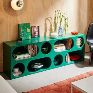May include: A bright green storage unit with oval openings, filled with books, bowls, and a wire basket. The unit is against a light brown wall, with a red rug and a white table in front. The unit is approximately 150 cm long.