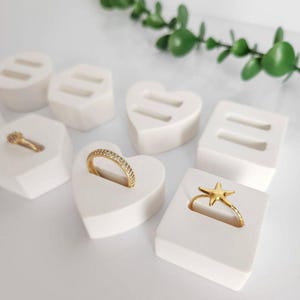 Op de afbeelding: Gouden ringen op witte ringhouders. De ringen hebben verschillende ontwerpen, waaronder een band met kleine steentjes en een zeester. De houders zijn hart- en vierkant van vorm. Groen gebladerte op de achtergrond.