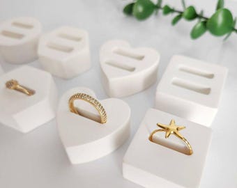 May include: A collection of gold rings displayed on white ring holders. The rings feature various designs, including a band with small stones and a starfish. The holders are in heart and square shapes.