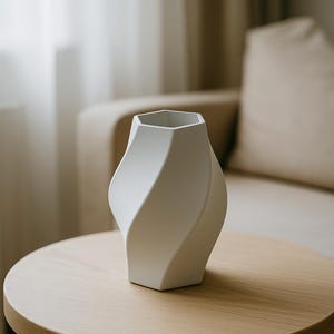 May include: A white, modern vase with a unique, twisted design. The vase has a hexagonal top and a curved, flowing shape. It sits on a round, light-colored wooden table. The background includes a beige sofa and curtains.