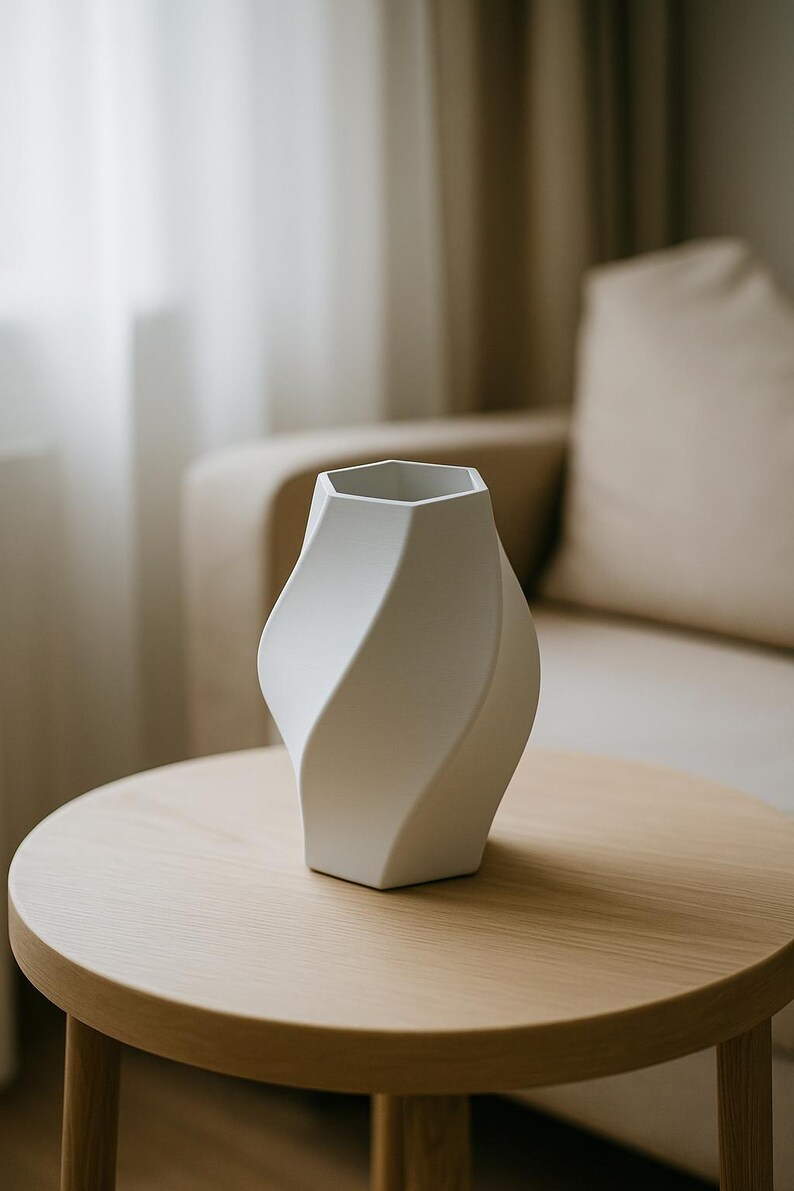 May include: A white, modern vase with a unique, twisted design. The vase has a hexagonal top and a curved, flowing shape. It sits on a round, light-colored wooden table. The background includes a beige sofa and curtains.