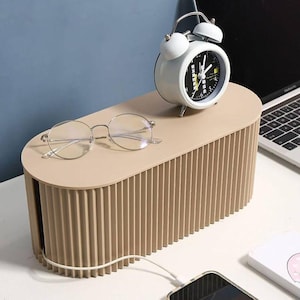 Op de afbeelding: Een beige kabelorganizer met een geribbeld ontwerp, met een witte wekker en een ronde bril. Een smartphone wordt opgeladen en een laptop is zichtbaar op de achtergrond. De organizer is ontworpen om kabels te verbergen en te beheren.